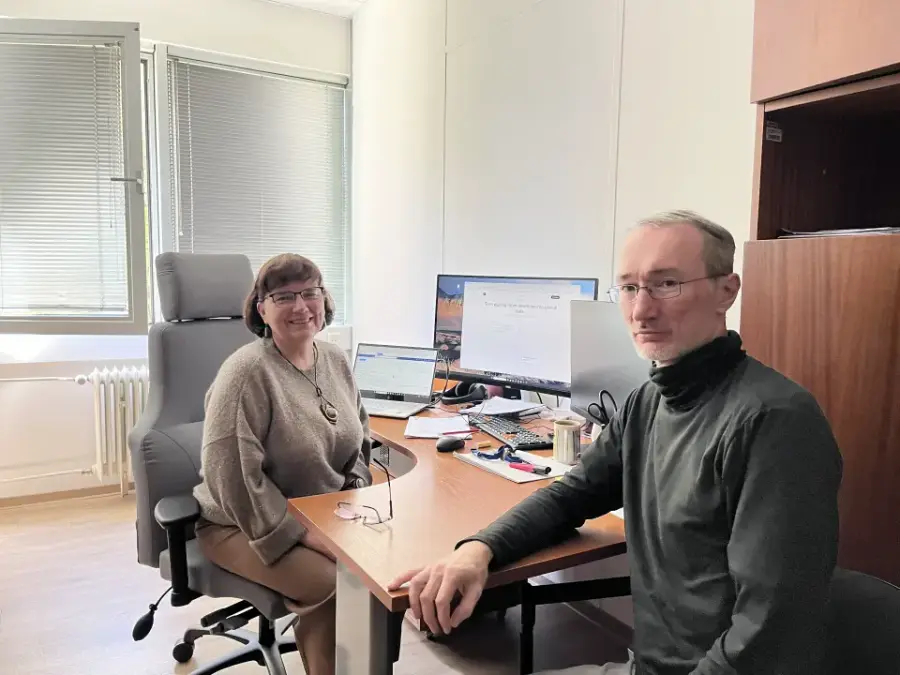 Two researchers at a shared office desk, a monitor and laptop showing project material between them.