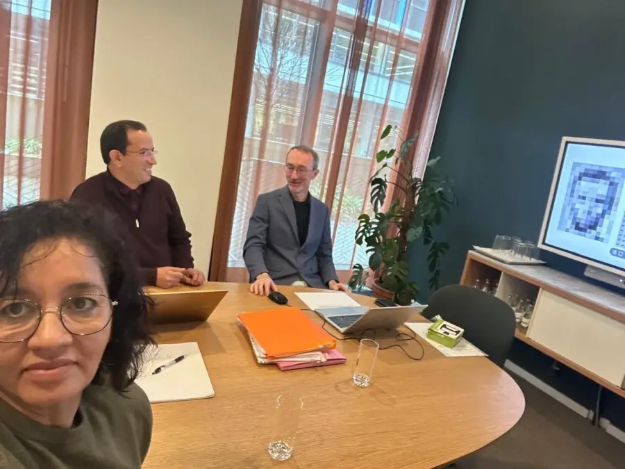 Project partners in conversation across a meeting table, analysis imagery displayed on the screen behind them.